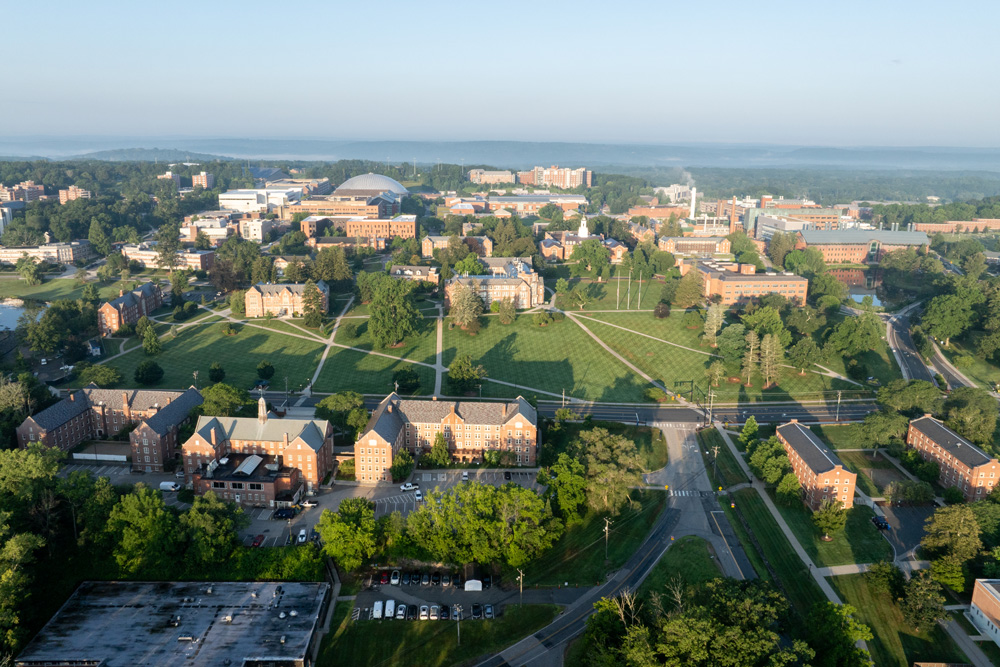 2025-07-25_StorrsSummerSunrise-42 Aerial view of a large university campus with academic and residential buildings, green lawns, tree-lined paths, and a stadium in the distance under a clear blue sky.