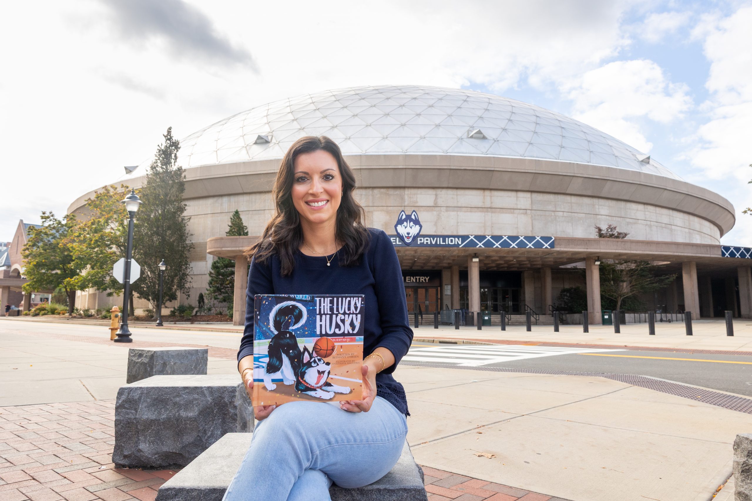 Bethany O'Brien '11 (CLAS) sits near Gampel Pavilion with a copy of her children's book, "The Lucky Husky," on Friday, Sept. 26, 2025. (Sydney Herdle/UConn Photo)