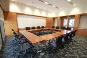 Photo of long wooden tables arranged in a square. There are four rolling chairs lined up on each side.