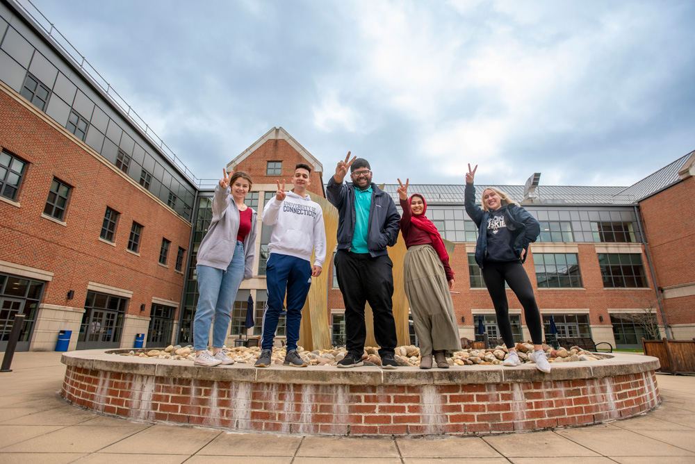 Group of students standing and jumping at the fountain in the outdoor courtyard at the UConn Waterbury campus.