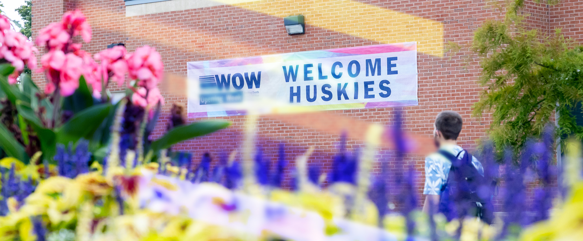 Welcome back Huskies banner hangs on a brick building with a person walking and flowers in the foreground.