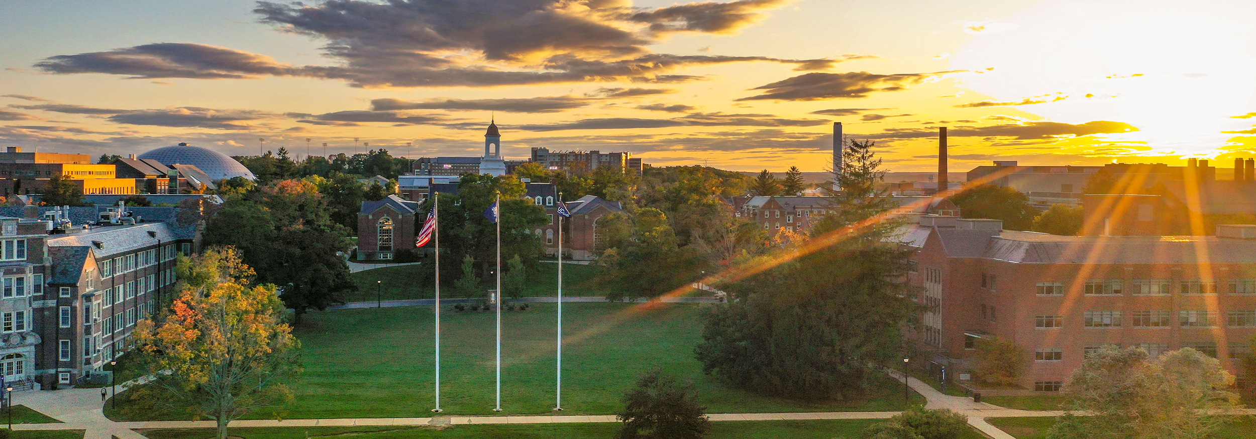 UConn Storrs campus at sunset with academic buildings, flagpoles, autumn trees, and golden evening light.